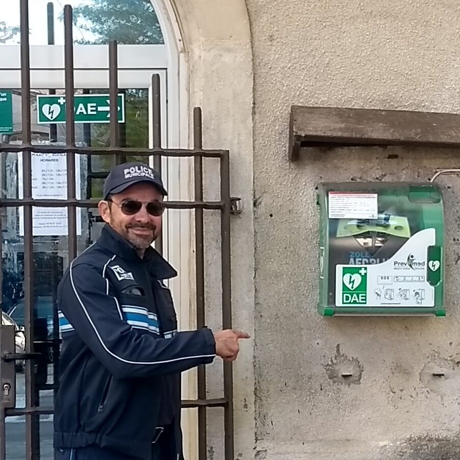 Photo de l'entrée du poste de Police municipale avec un agent montrant l'emplacement du défibrillteur